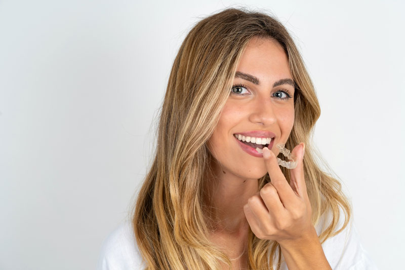 A smiling woman holding a clear aligner, preparing to place it over her teeth, showcasing the use of Invisalign for discreet orthodontic treatment.
