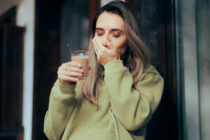 Woman in a green hoodie wincing in pain while holding a glass of cold beverage, covering her mouth, possibly due to tooth sensitivity.