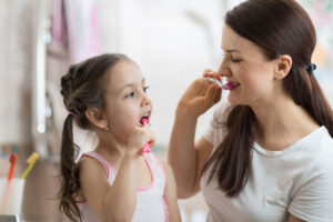 Mother and young daughter brushing their teeth together in a bright bathroom, demonstrating healthy oral hygiene habits for children.