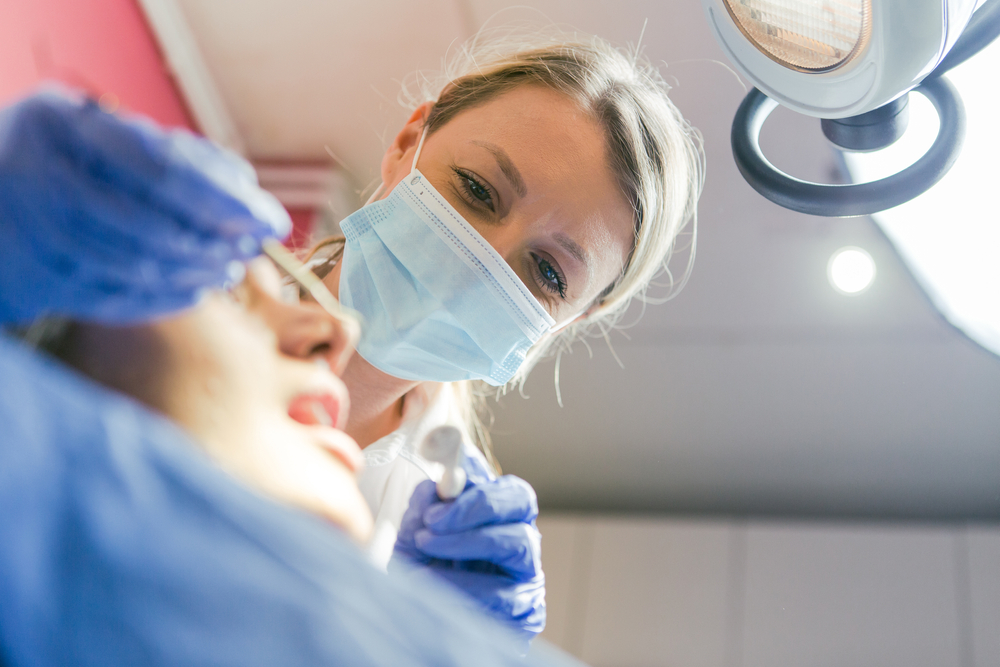 A dentist wearing a mask and gloves examines a patient’s mouth with dental tools under a bright examination light in a dental clinic.